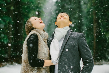 A man and woman in winter clothes look happily to the sky as the snow falls in the forest. 
