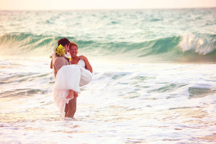 Man dressed in suit holding woman in wedding dress while standing in ocean