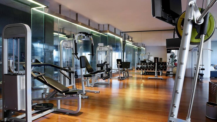 Spacious fitness room with hardwood floors, lit windowed walls, and numerous exercise machines and TVs. 