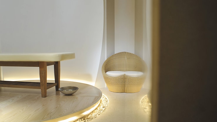 A softly lit white room with a wicker chair and a lit, raised platform holding a massage table.