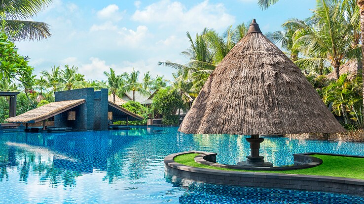 A crystal blue lagoon pool with in-water seating under a thatched umbrella, accessed by a synthetic sandbar. 