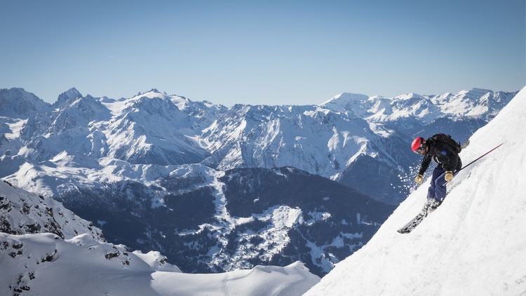 Person skiing down mountain side with snowy mountains in background.