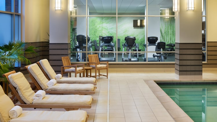 Cushioned lounge chairs bordering an indoor pool, with fitness center in background.