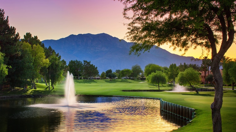A golf course with a pond and fountain and mountains in the distance.