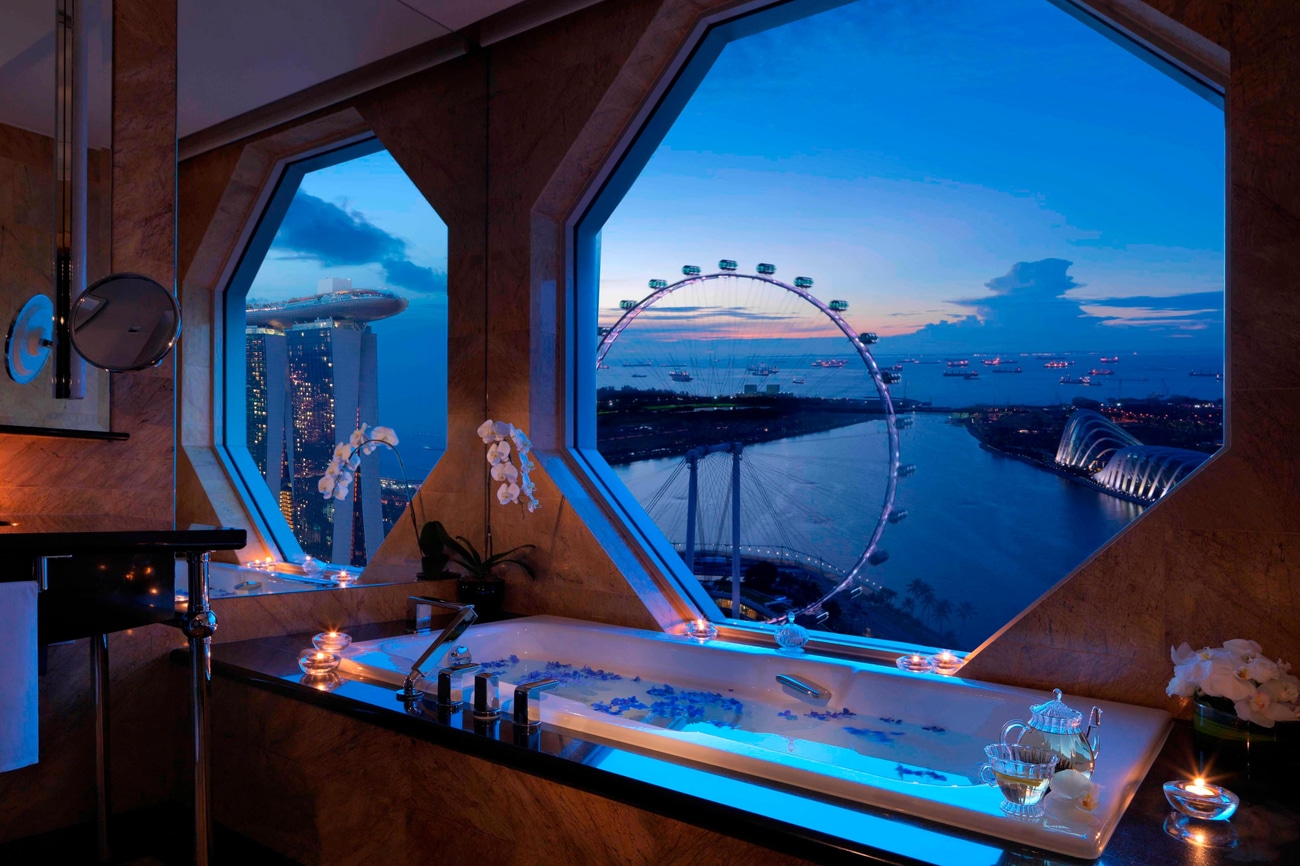 Marble bathroom with a bathtub and an octagonal window overlooking views of the Singapore Flyer