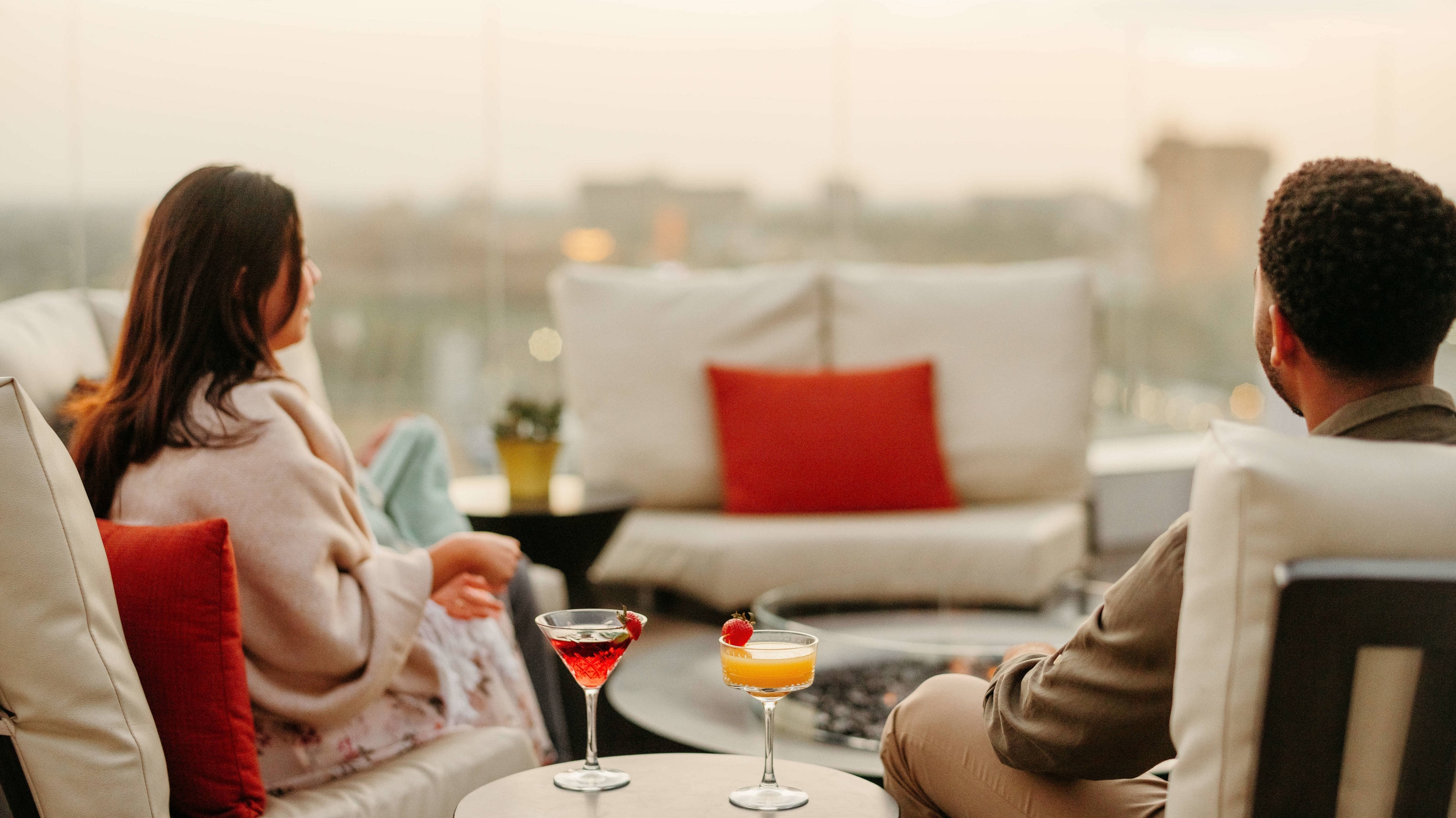 A couple sitting in chairs on a rooftop with cocktails served on the table between them.