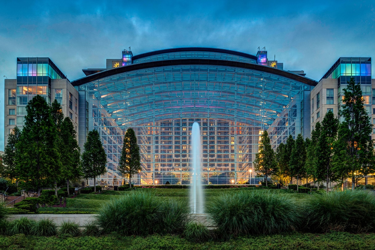 View of exterior of the atrium of the Gaylord National Resort & Convention Center, a tree-lined lawn and a fountain at sunset.