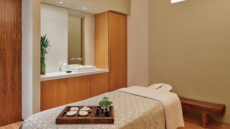 A spa bed with towels and candles sitting ready on it in a comfortably small room with nearby sink and mirror.