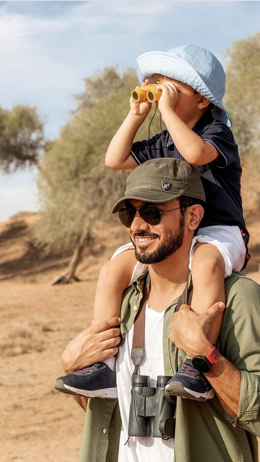 Father holding his son on his shoulders so the kid can look through binoculars for animal watching.