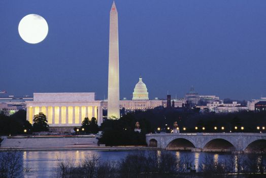 A full moon shines down on a columned building, domed building and obelisk across the water