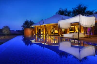 A tented, house-like structure and an outdoor bed with a canopy overlook a pool in the evening