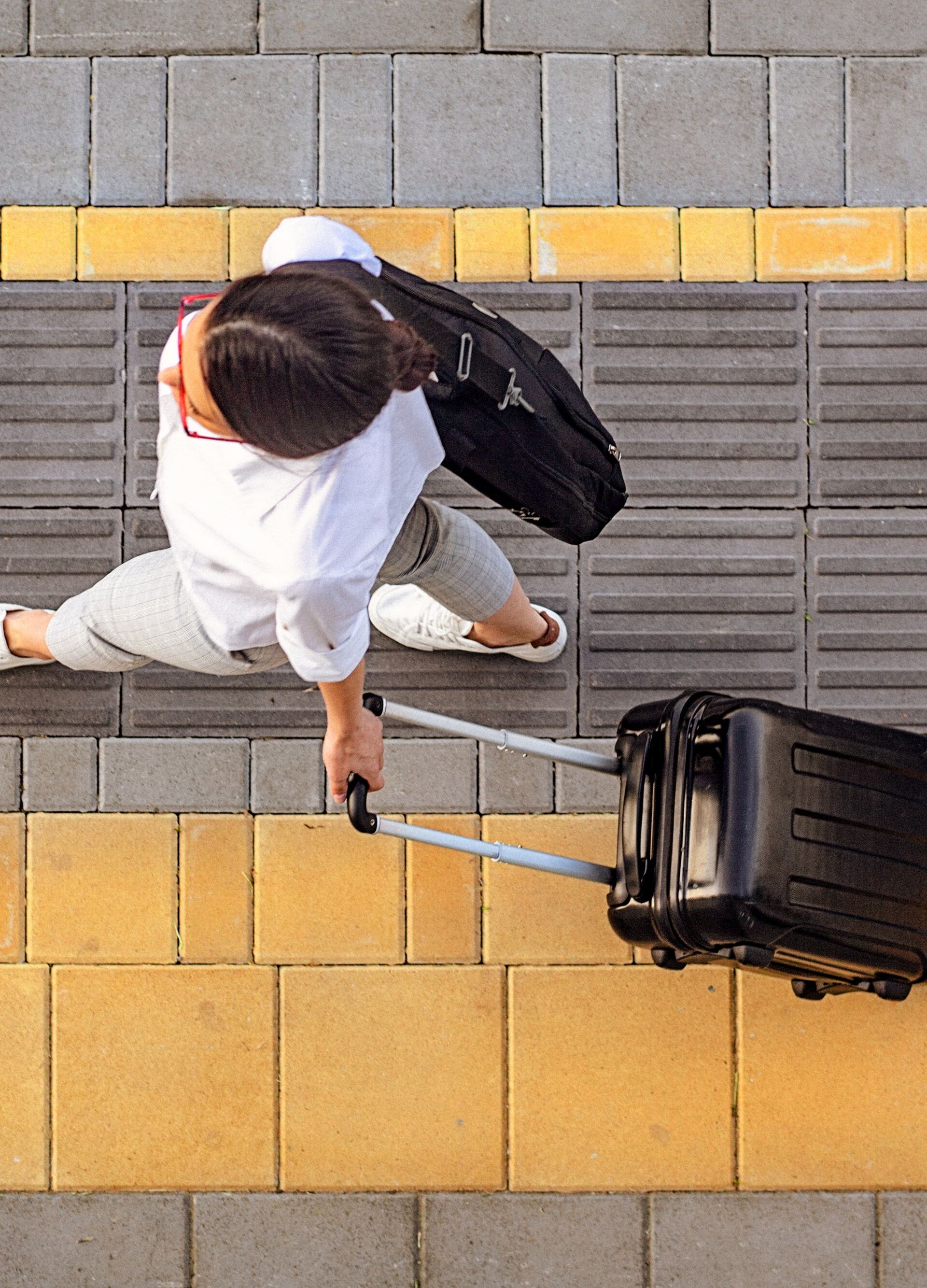 High angle view of a young woman walking on a sidewalk and pulling a small wheeled luggage with a briefcase on it