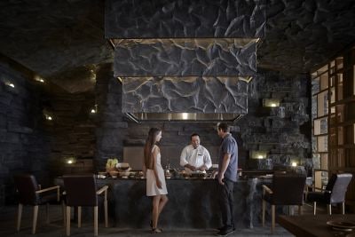 A man in chef?s whites stands behind a food-filled counter talking to a man and woman in a room with stone walls