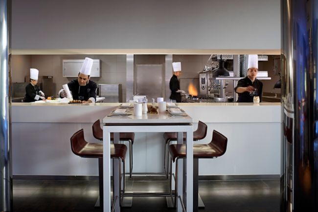 A high-top dining table with four stools in front of an open kitchen where chefs are preparing food