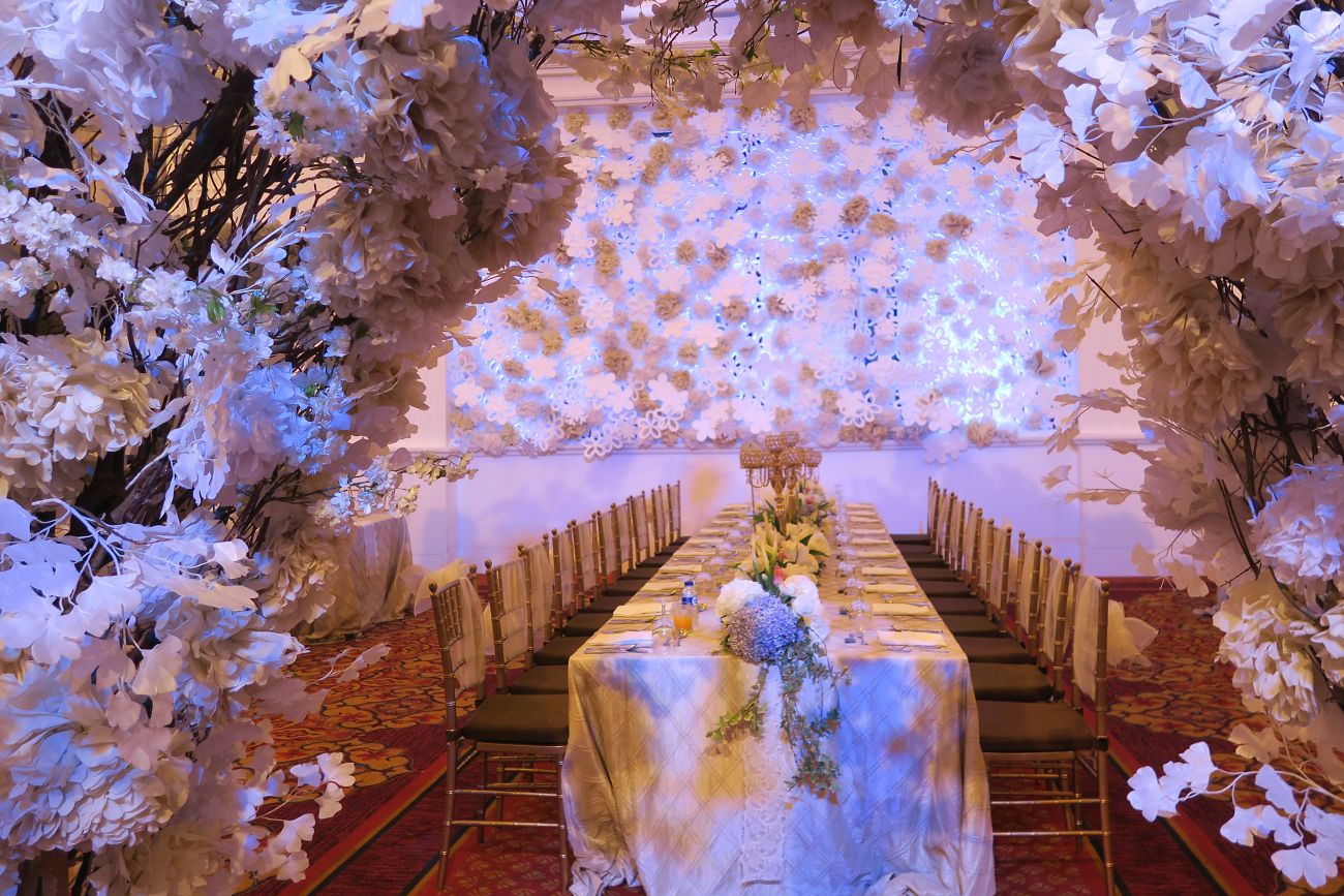 An archway of white flowers opens onto an event space with a long table draped in white featuring colorful florals and chairs