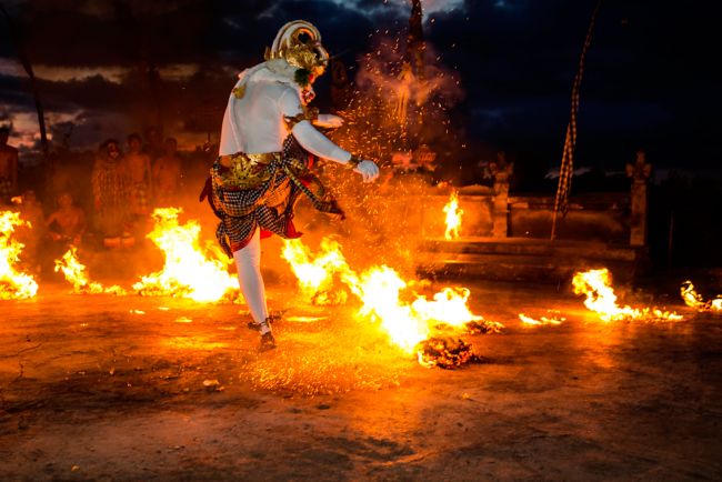 Man in a white body suit and ram-like mask dances with fire in front of a nighttime crowd