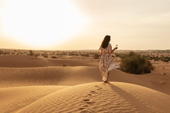 امرأة تمشي وسط الكثبان الرملية وهي تحمل مشروبًا