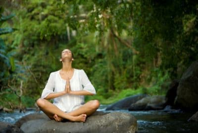 Woman in a white tunic sits cross-legged on a boulder whilst meditating in the rainforest