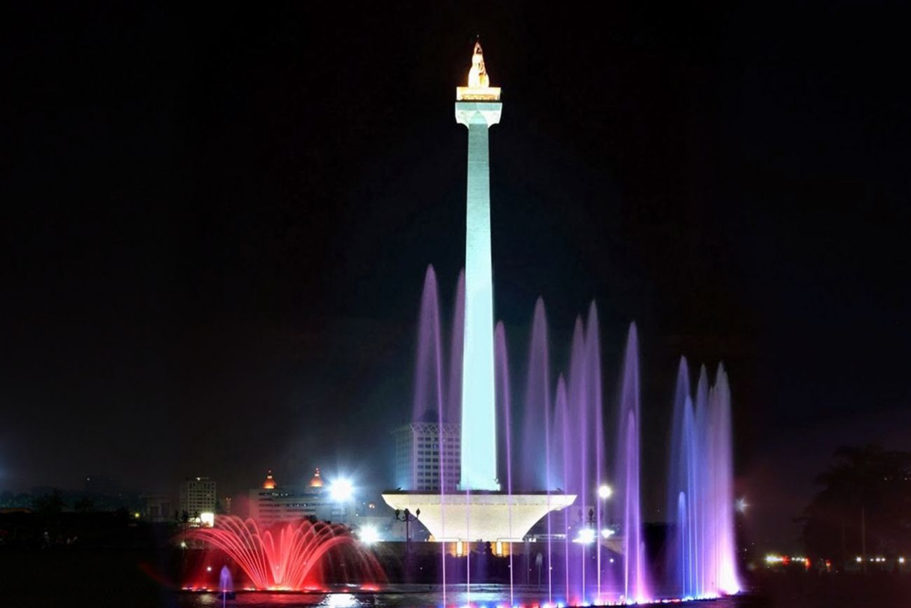 Fountains illuminated in purple and red surround the soaring obelisk of the National Monument at night