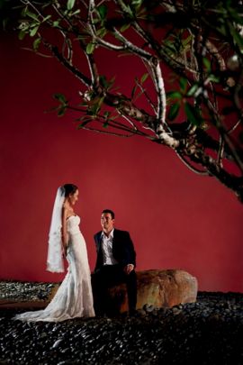 Bride stands over a groom sitting on a boulder against a red wall at night as they smile at one another