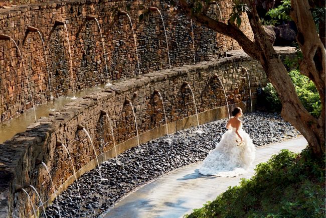 Bride in a strapless white gown with a full skirt glides by a stacked stone wall with water spouts