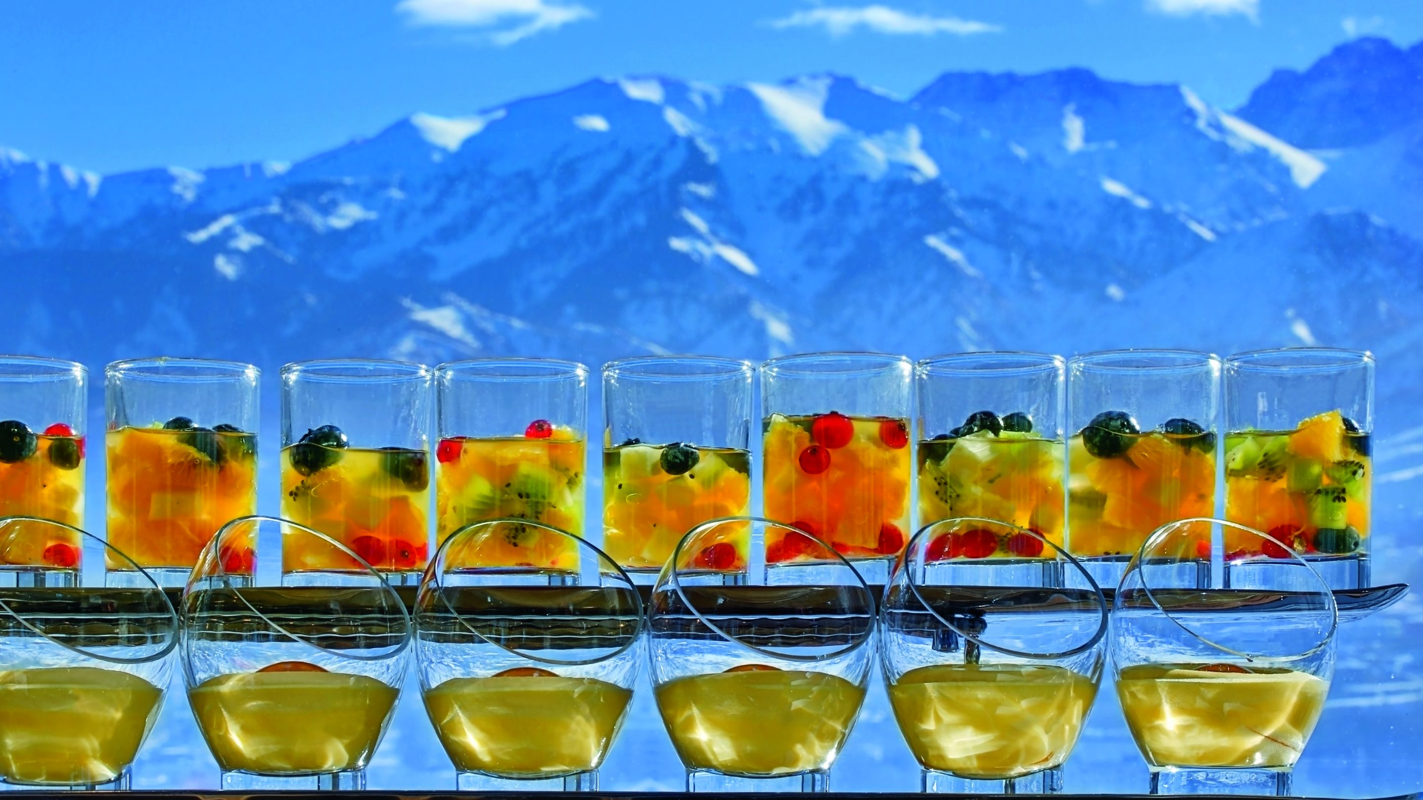 A row of glasses, filled with fruit cocktails and custard, overlooking snow-capped mountains