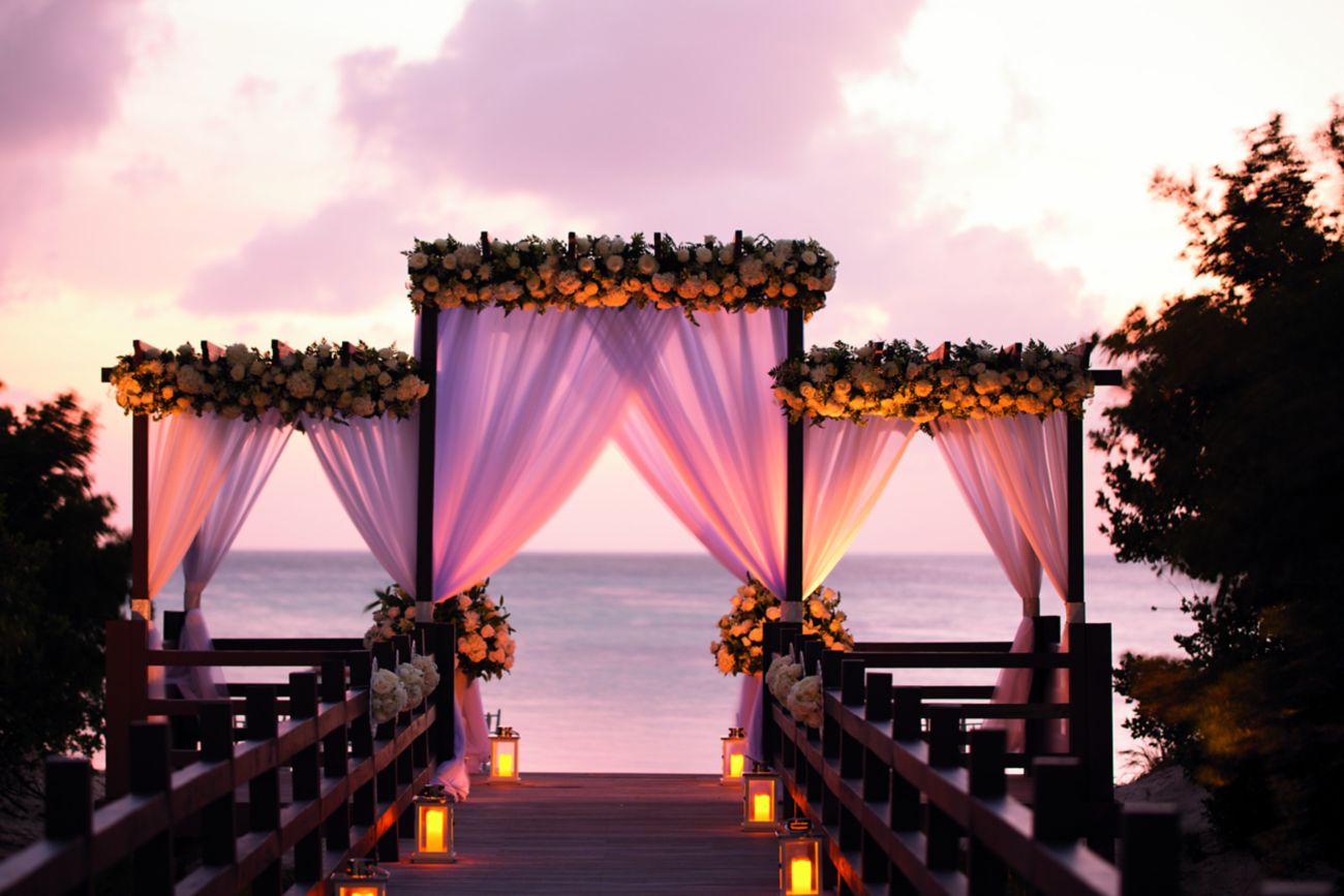 A candle-lined pier leads to a gazebo adorned with flowers and fabric overlooking the ocean at sunset