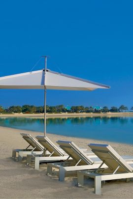 Four white lounge chairs and an umbrella on the beach overlooking a quiet lagoon