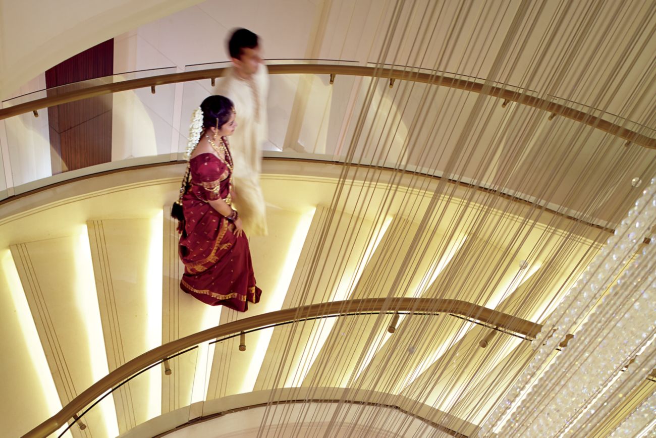 Couple in traditional Indian wedding attire walk along a curvaceous staircase suspended next to a crystal light fixture