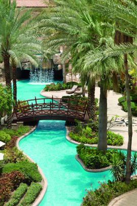 Lazy river feature with a bridge and waterfall, all of which is hemmed in by landscaping and lounge chairs