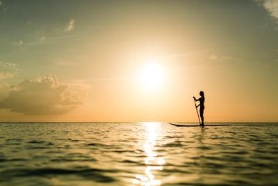 A woman holding a pole stands on a surfboard in the ocean at sunset