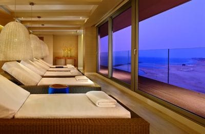 A row of woven chaise lounges with plush white cushions line up in front of floor-to-ceiling windows overlooking the sea