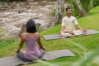 A man and woman face each other on a green lawn next to the river while meditating