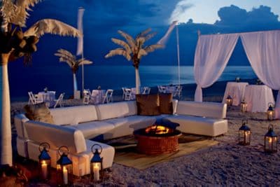 Hurricane lamps illuminate white couches, palm trees and drapes during an evening reception on the beach