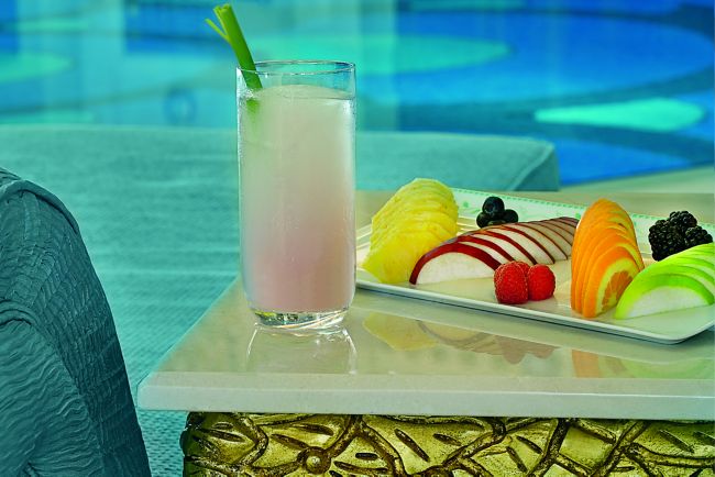 A light pink drink and a platter of fresh, colorful fruit rest on top of a white table next to the pool