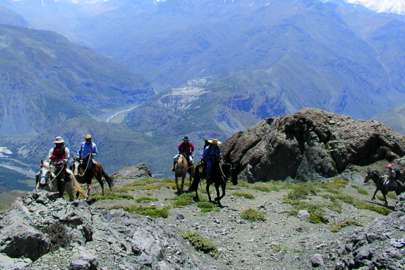 People on horseback stop at a ledge to look out over a vast mountain valley