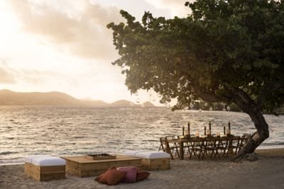 A long dining table and seating area set up near a large tree at the ocean?s edge