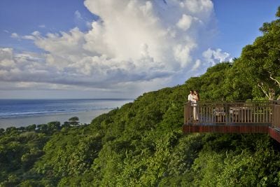 A man and woman standing on a wooden balcony overlooking lush greenery and the ocean