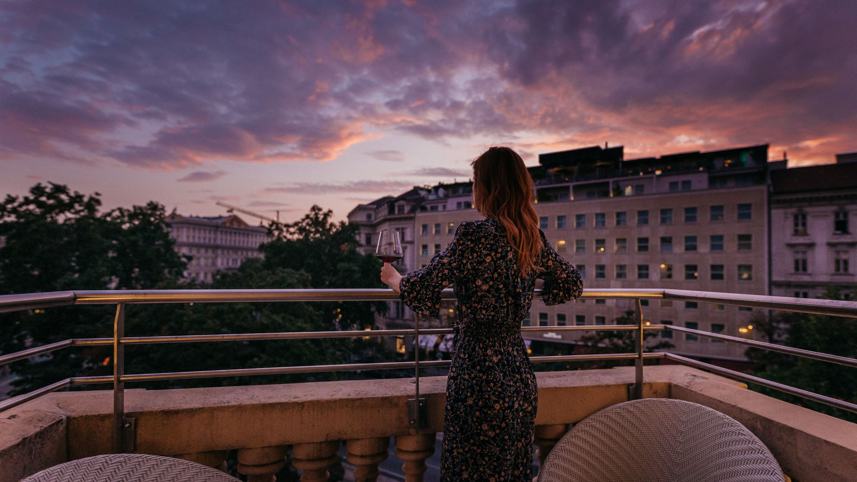 A woman standing on a balcony, enjoying the city view and a scenic sunset.