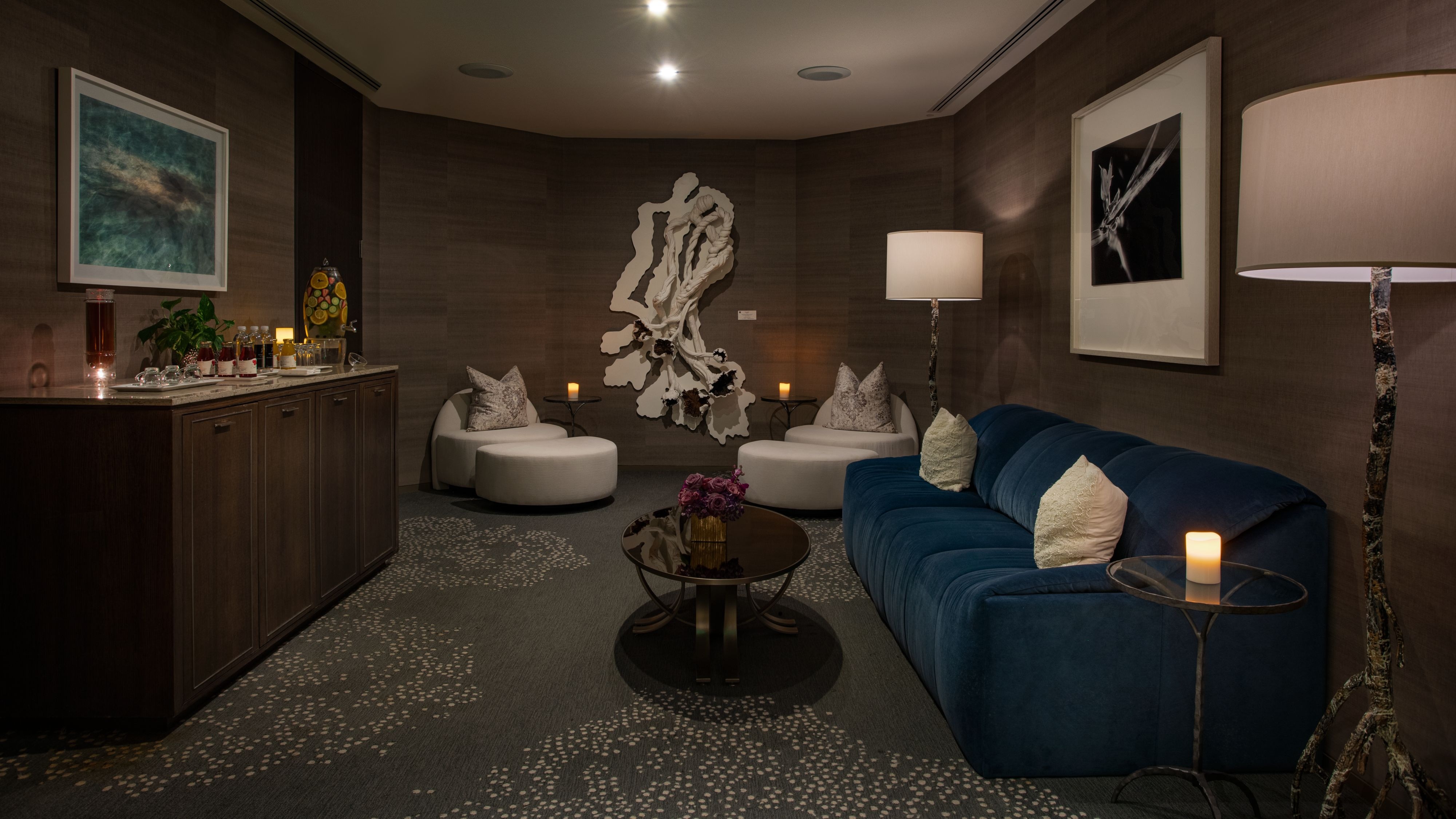 Dim lounge with dark textured walls, a long navy sofa, a low table with flowers, white ottomans, and a wall sculpture.