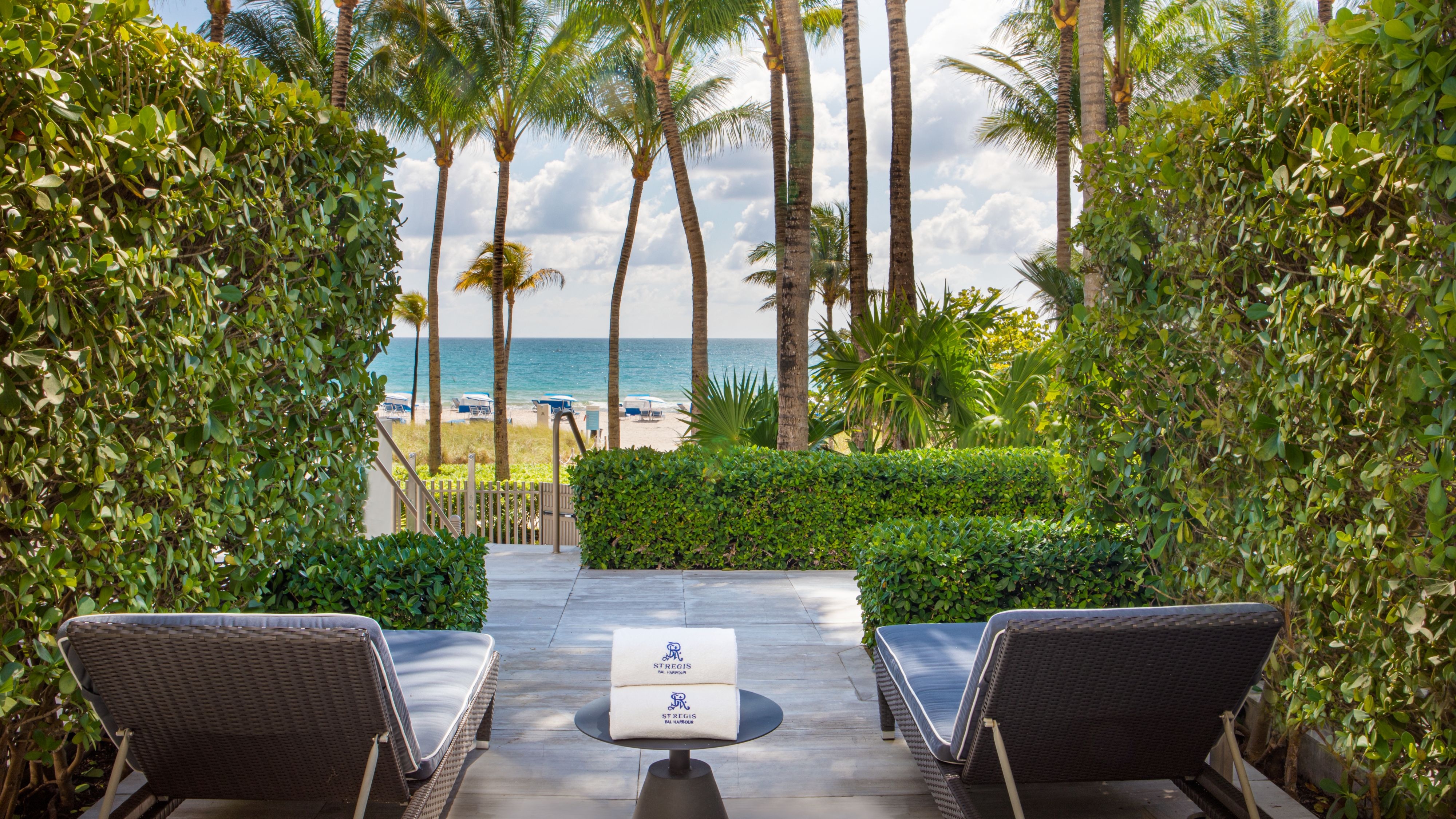 Paved terrace between two tall hedges, with two dark loungers, a small table holding two rolled towels, and palms near the sea.