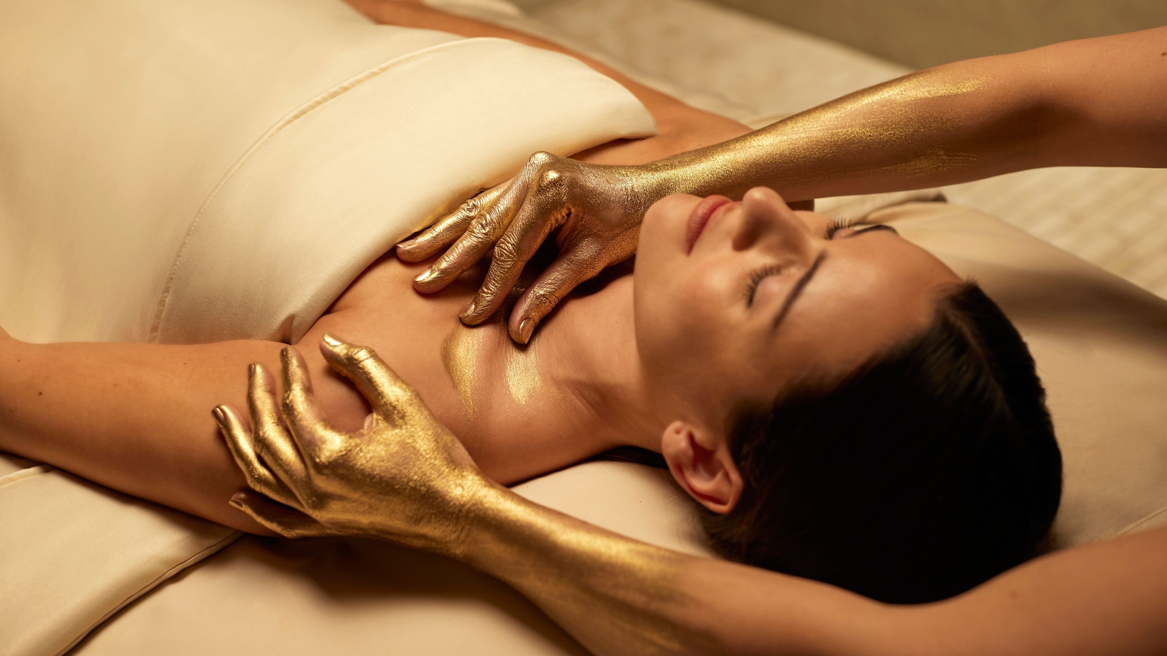 A woman lies back in The Spa at The St. Regis Bal Harbour with a towel across her chest and two gold-painted hands on her shoulder.