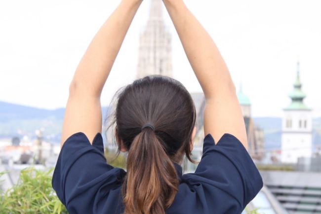 Woman in a navy t-shirt as seen from behind as she raises her hands above her head and faces the skyline