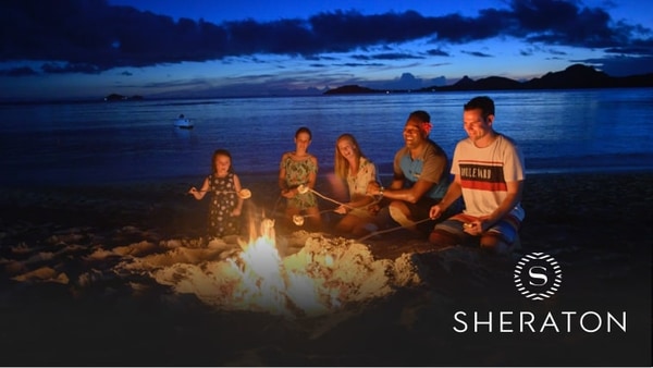 Five people roasting marshmallows around a fire on the beach with a boat in the water beyond