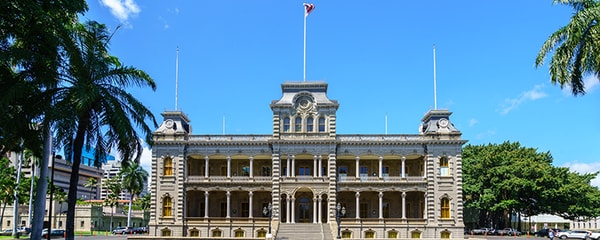 Full view of Iolani Palace in Hawaii on a sunny day surrounded by green trees.